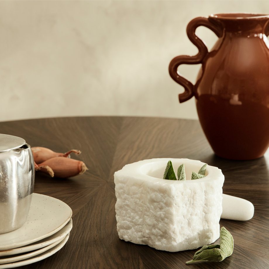 Ferm Living Petra Mortar, een elegante witte kwarts vijzel met stamper, geplaatst op een donkere houten tafel, omgeven door servies en een decoratieve vaas