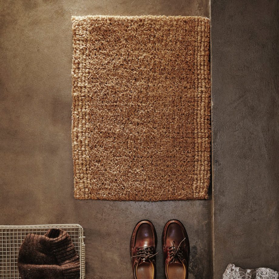 Ferm Living Coir Mat geplaatst op een betonnen vloer naast schoenen en een metalen mand met een sjaal. De natuurlijke kokosvezelmat heeft een lusvormige rand en voegt een rustieke, moderne stijl toe aan de entree