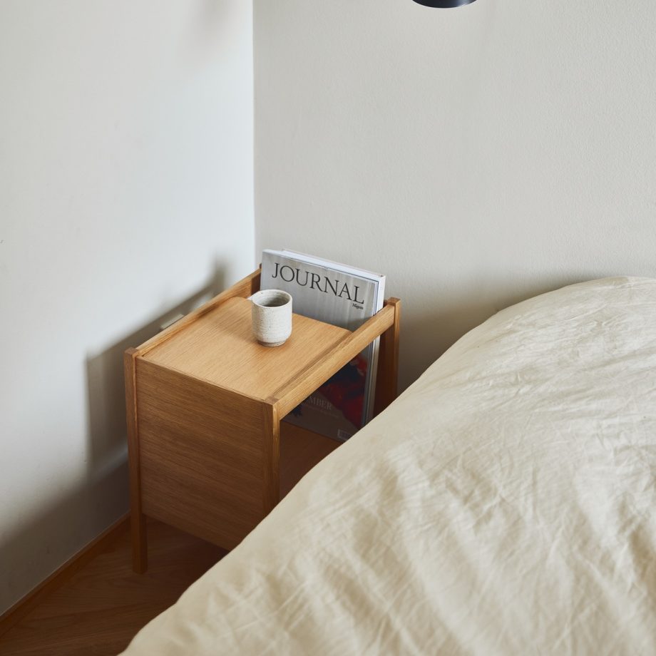 Form Refine Journal bijzettafel in eikenhout als nachttafel naast het bed met tijdschrift, kaars en een minimalistische lamp aan de muur voor een strakke, moderne uitstraling in de slaapkamer.