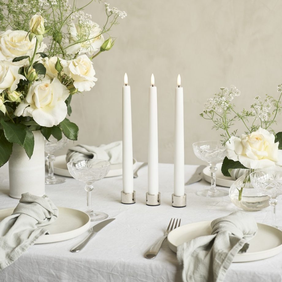 Drie witte LED-dinerkaarsen van Uyuni Lighting in chromen kandelaars, geplaatst op gedekte tafel met witte vasen met lichte bloemen en servies bij bruiloft