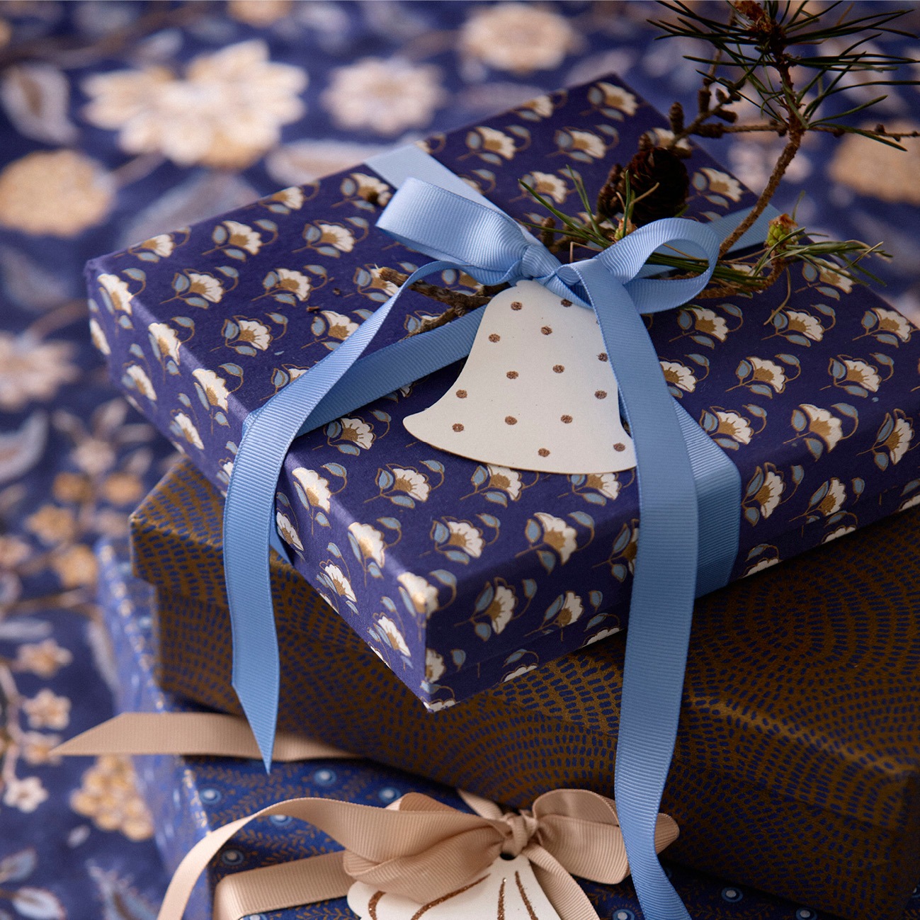 Luxe ingepakte cadeaus met blauwe en gouden details, versierd met Bungalow gift tags in de vorm van kerstbellen en strikken, geplaatst op een bijpassend patroonpapier.