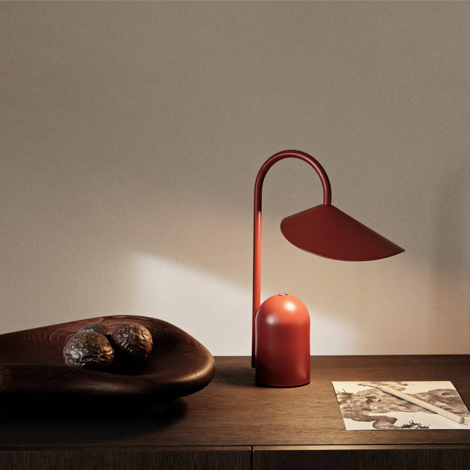 De Ferm Living Arum Portable Lamp in een rode uitvoering, geplaatst op een houten dressoir naast decoratieve elementen, verlicht sfeervol de omgeving