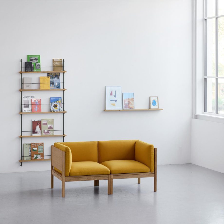 Modulaire sofa van Moebe met warmgele bekleding en eikenhouten frame, geplaatst in een minimalistisch Scandinavisch interieur met wandplanken en tijdschriftenrek.