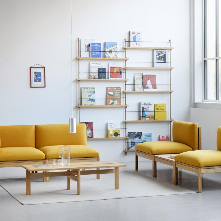 Een stijlvolle Moebe Modular Sofa in een zonnige gele tint, gecombineerd met een houten salontafel en een modulaire wandkast gevuld met designboeken. Dit Scandinavische designmeubel biedt flexibiliteit met losse zitmodules, perfect voor een moderne woonkamer of lounge.