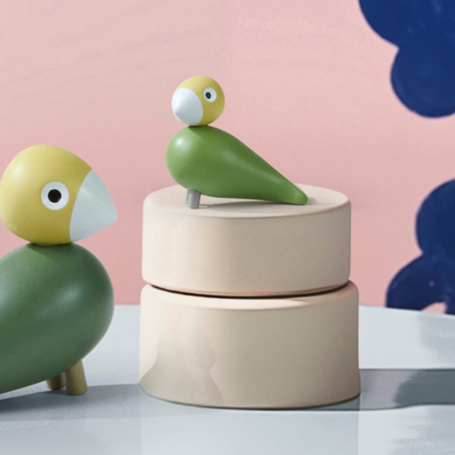 Set van twee Kay Bojesen Songbirds, Liv & Lykke, in zachte groene tinten. Handgemaakt van hout en geplaatst op een elegante beige doos tegen een artistieke roze en blauwe achtergrond. Perfecte Scandinavische decoratie voor liefhebbers van design en natuur.