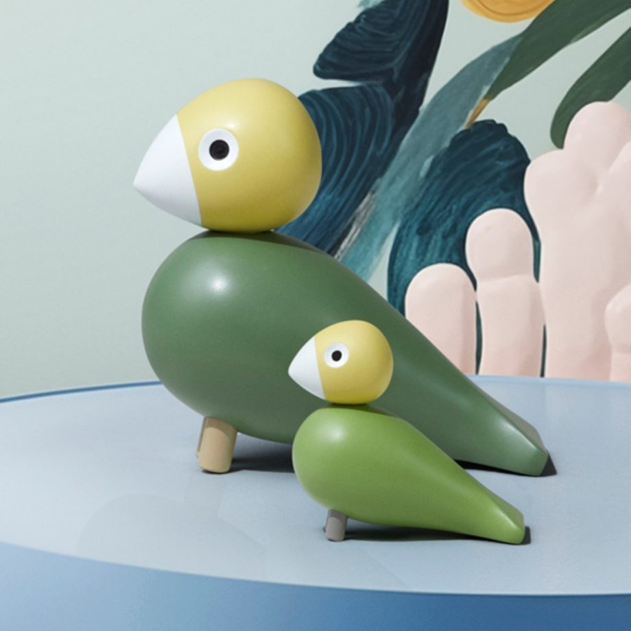 Set van twee Kay Bojesen Songbirds, Liv & Lykke, in groene tinten, elegant gepresenteerd op een blauwe tafel tegen een kleurrijke kunstachtergrond. Tijdloos Scandinavisch design, handgeschilderd in hout en perfect als interieurdecoratie of cadeau