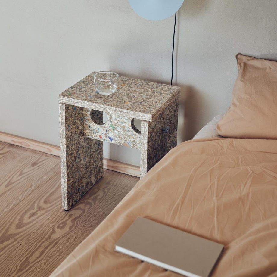 Nachtkastje gemaakt van gerecycled materiaal naast een bed met beige dekbedovertrek, met wandlamp en glas van Form and Refine.