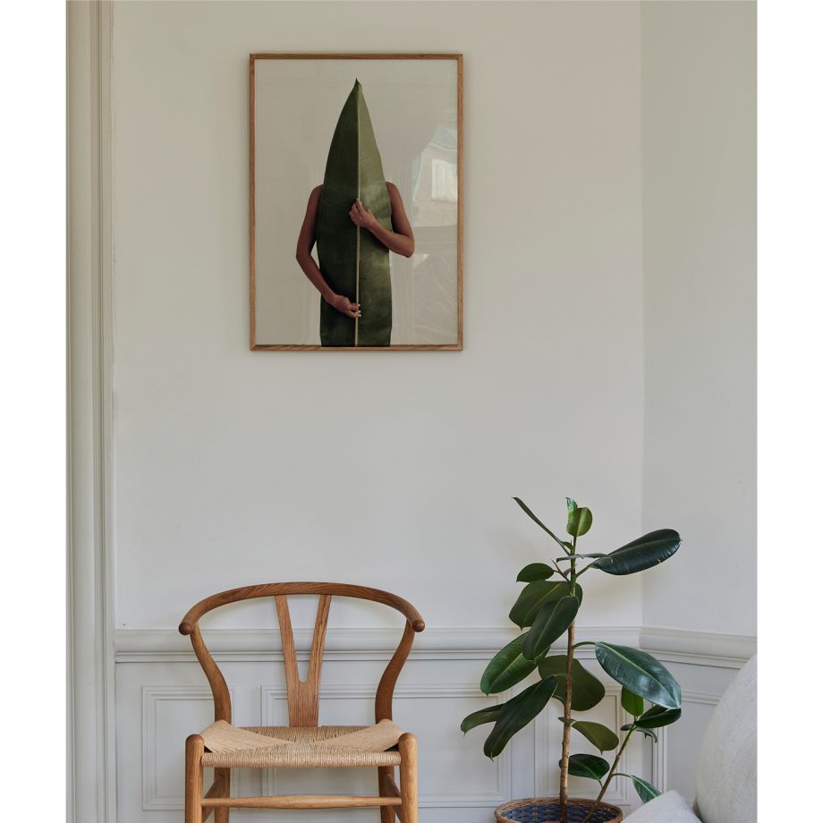 Paper Collective Naturaleza poster van figuur achter een groot groen blad hangend boven een scandinavische stool